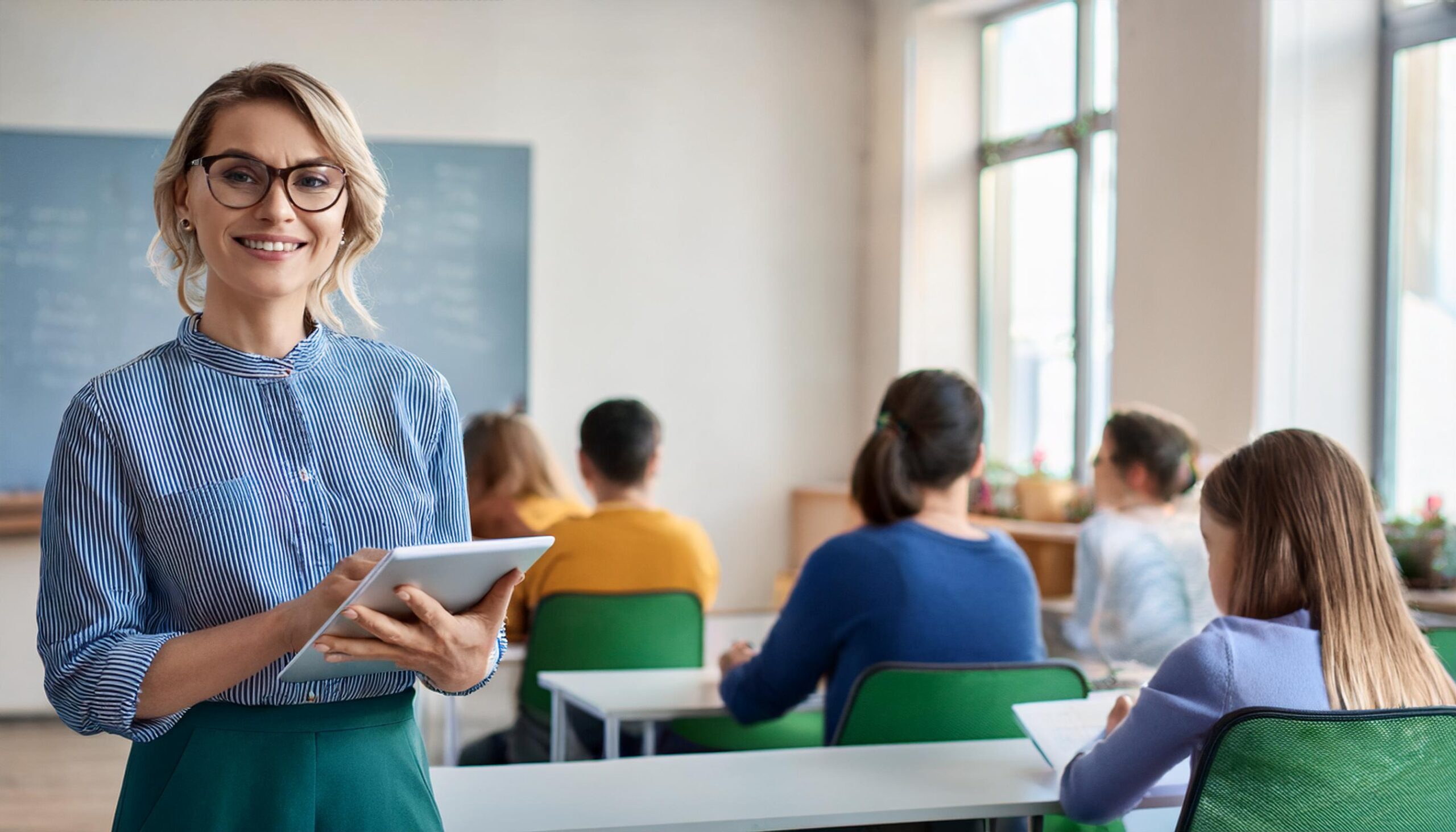 Klassenlehrerin steht im Vordergrund mit einem Tablet in den Händen und ich Hintergrund sitzen die Schüler an ihren Schreibtischen.