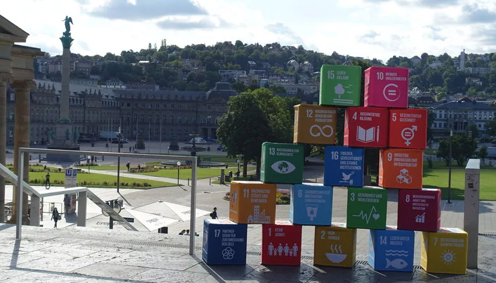 17 verschiedenfarbige Quader stehen zu einer Pyramide aufgetürmt auf der Treppe oberhalb des Schlossplatzs in Stuttgart, mit Blick in Richtung Neues Schloss. Auf den Blöcken stehen die 17 Ziele globaler Nachhaltigkeit der Vereinten Nationen.